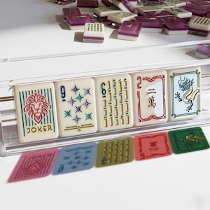May include: A collection of ivory-colored Mahjong tiles with various symbols and numbers, including a lion, stars, and Chinese characters. The tiles are arranged in a clear acrylic holder, with additional tiles scattered in the background. The word "JOKER" is visible.