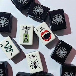 Op de afbeelding: Een verzameling rechthoekige speelstenen met zwarte stenen met witte spinnenwebpatronen en witte stenen met diverse illustraties, waaronder een groene gifvles, een rode mond met hoektanden, een groene slang en een zwarte spin.