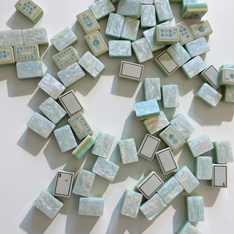 May include: A collection of rectangular mahjong tiles featuring a pearlescent light blue and white marbled finish. The tiles display various engraved symbols, numbers, and blank white faces with thin borders, scattered across a plain surface.