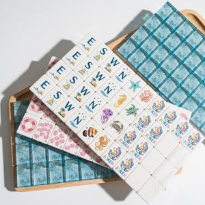 May include: A set of white mahjong tiles featuring colorful illustrations of sea creatures, mermaids, and nautical symbols, alongside blue translucent tiles. The tiles display letters E, S, W, N, and the word JOKER, arranged on a wooden tray.