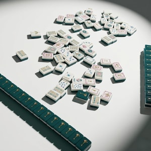 May include: A collection of white and teal Mahjong tiles scattered on a white surface. The tiles feature various symbols and designs in gold, teal, and pink. A teal rack with gold accents is visible on the left and right sides of the image.