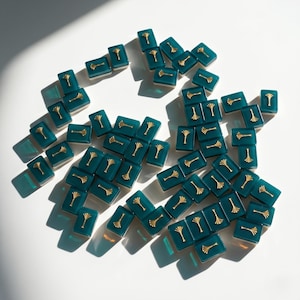 May include: A collection of teal rectangular game tiles with gold-colored tree designs. The tiles are scattered on a white surface, with shadows adding depth. The gold accents provide a contrast to the teal color.