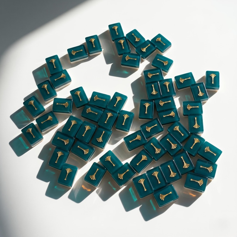 May include: A collection of teal rectangular game tiles with gold-colored tree designs. The tiles are scattered on a white surface, with shadows adding depth. The gold accents provide a contrast to the teal color.