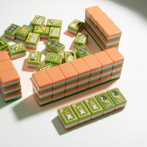 May include: A set of mahjong tiles featuring a three-layered design with a peach-colored top, a white middle, and a green base. The tiles display various green-toned illustrations, Chinese characters, and English text including West, East, and Joker.