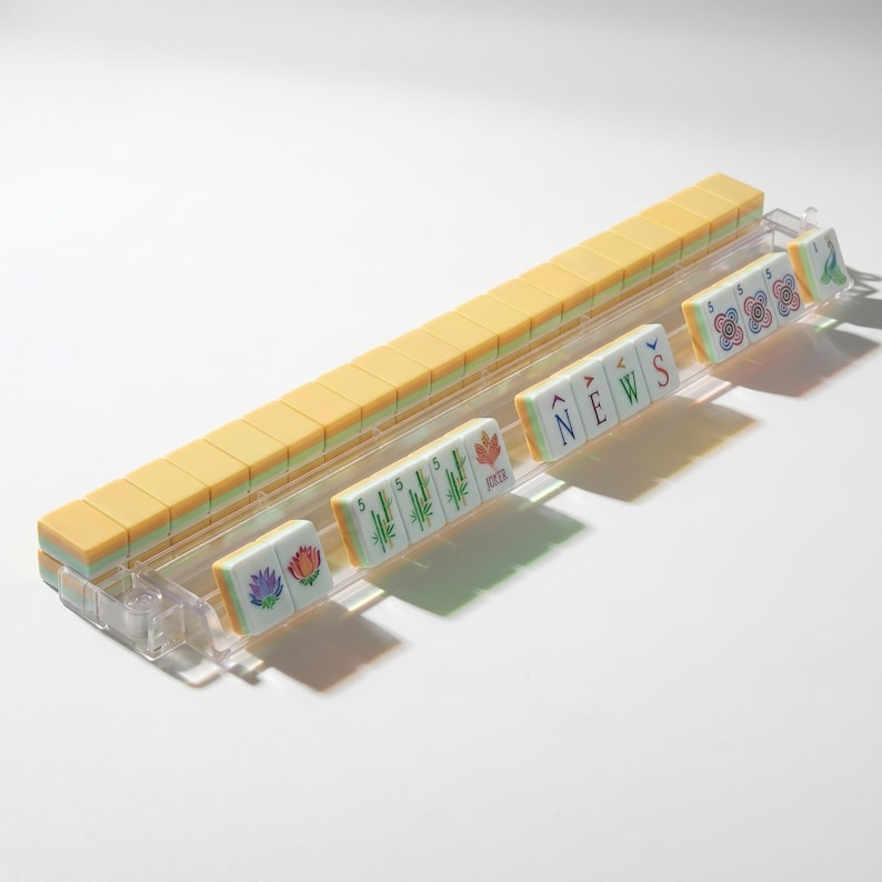 May include: A clear plastic Mahjong rack holding a row of yellow-backed tiles. The front-facing tiles display colorful floral designs, green bamboo stalks, the word NEWS in blue and red, and the number 5 with circular patterns in red and blue.