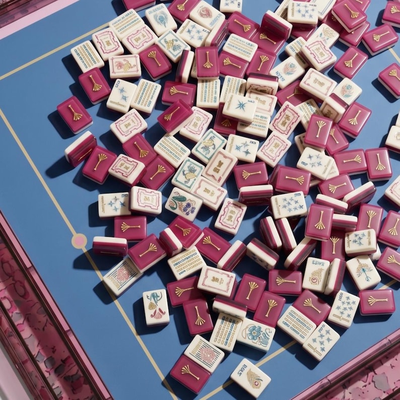 May include: A collection of mahjong tiles in shades of burgundy and white, scattered on a blue surface. The tiles feature various symbols and designs, with gold accents. The game pieces are arranged on a blue game board with pink trim.