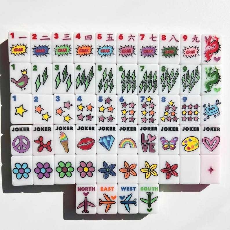 May include: A set of white rectangular mahjong tiles featuring colorful printed graphics including numbers, Chinese characters, lightning bolts, stars, flowers, hearts, rainbows, diamonds, lips, airplanes, and the word JOKER on specific tiles.