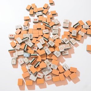 May include: A collection of rectangular mahjong tiles scattered across a surface. The tiles feature a solid peach-colored back and a white front face with various black, green, and red engraved symbols, characters, and illustrations.