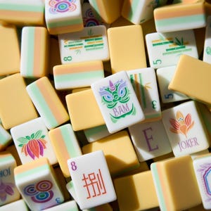 May include: A collection of rectangular mahjong tiles featuring a mix of cream, pale green, and soft orange colors. The tiles display various engraved designs including bamboo stalks, floral patterns, the word BAM, the word JOKER, and the letter E.