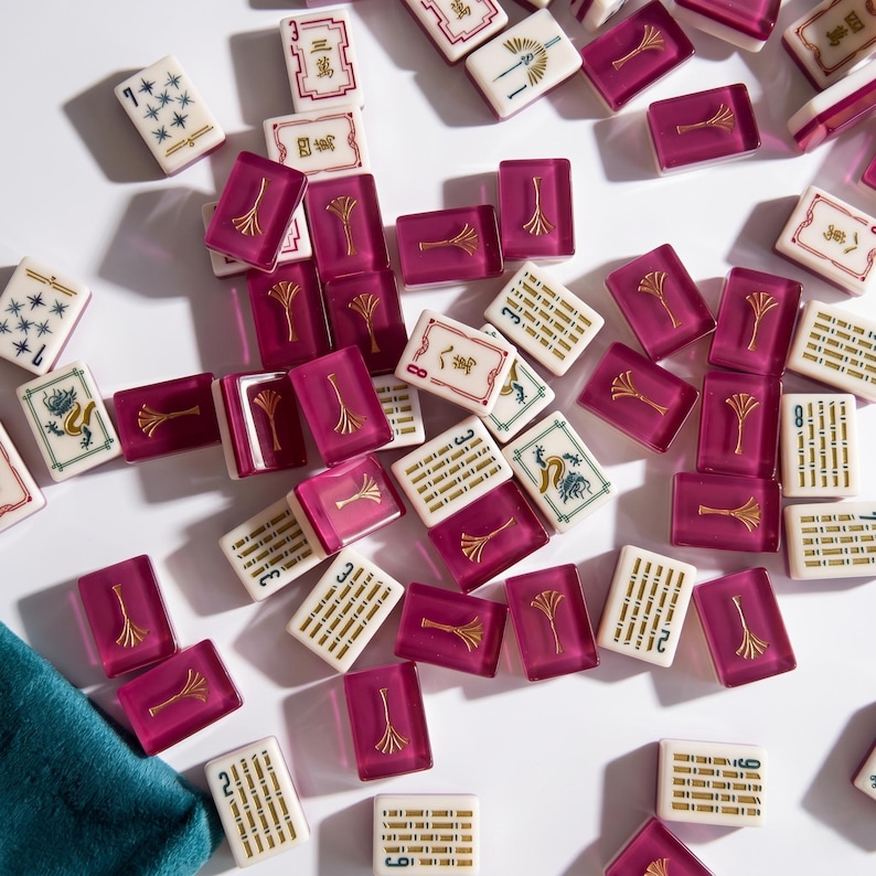 May include: A collection of mahjong tiles scattered on a white surface. The tiles feature a mix of colors, including white, magenta, and gold. Some tiles have symbols and numbers, while others have decorative designs. A teal cloth is visible in the lower left corner.