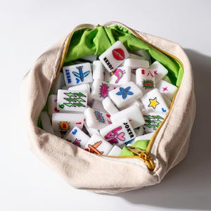 May include: A beige canvas zippered pouch filled with white rectangular mahjong tiles. The tiles feature colorful graphic illustrations including a blue airplane, red lips, a pink bird, a blue flower, a yellow star, and text reading JOKER and LOVE.
