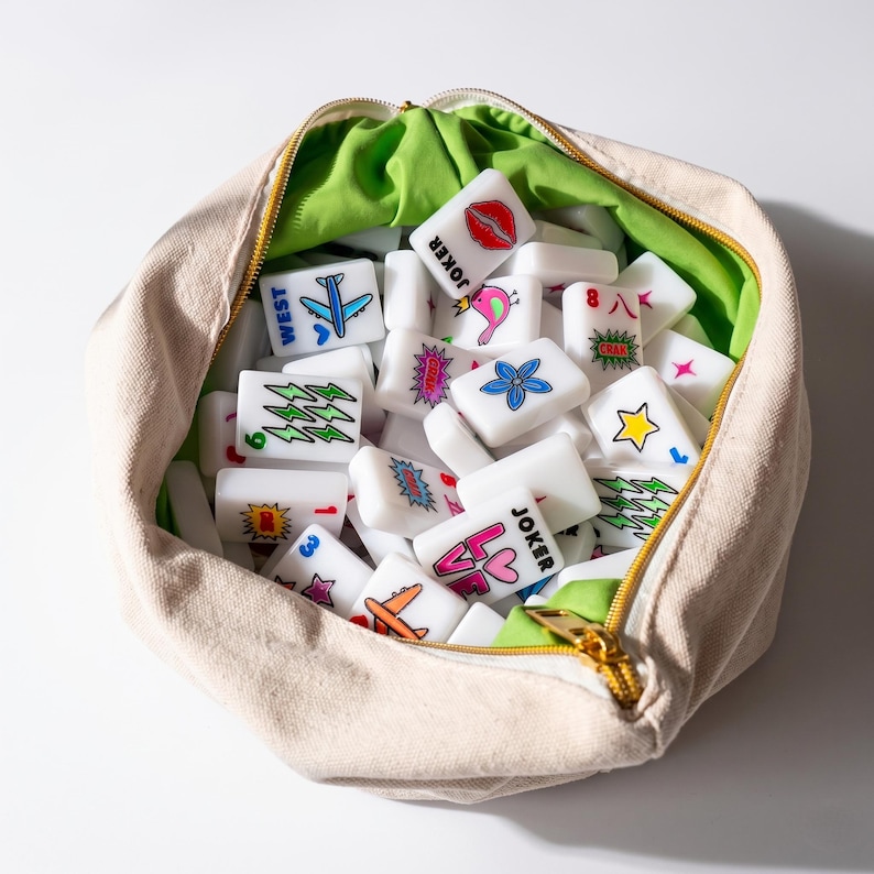 May include: A beige canvas zippered pouch filled with white rectangular mahjong tiles. The tiles feature colorful graphic illustrations including a blue airplane, red lips, a pink bird, a blue flower, a yellow star, and text reading JOKER and LOVE.