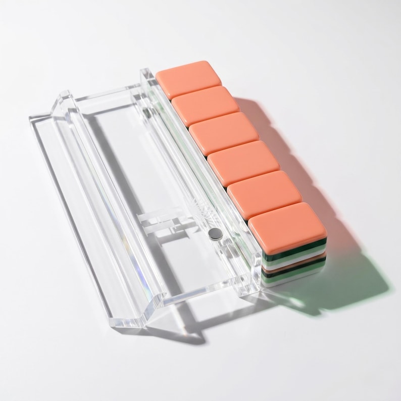 May include: A clear acrylic rack holding a stack of five rectangular tiles. Each tile features a solid peach-colored top surface with layered green, white, and dark green edges. The rack is transparent with a smooth, angled design and a small silver fastener.
