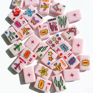 May include: A collection of light pink rectangular mahjong tiles featuring colorful printed graphics including airplanes, stars, lightning bolts, flowers, birds, butterflies, soap, and text such as JOKER, LOVE, NORTH, EAST, and CRAK.
