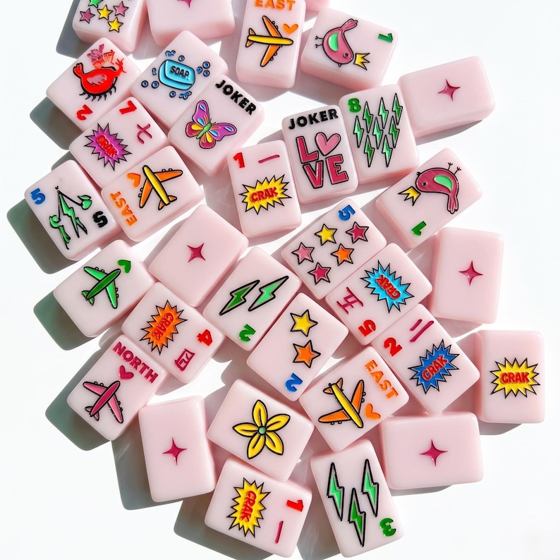 May include: A collection of light pink rectangular mahjong tiles featuring colorful printed graphics including airplanes, stars, lightning bolts, flowers, birds, butterflies, soap, and text such as JOKER, LOVE, NORTH, EAST, and CRAK.
