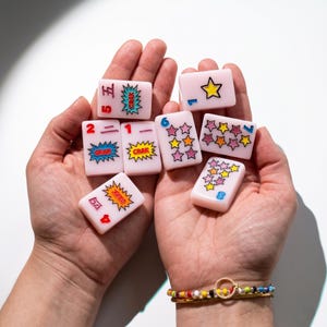 May include: A collection of seven pale pink rectangular mahjong tiles featuring colorful graphics. The tiles display various numbers, star patterns, and the text "CRAK" inside comic-style speech bubbles. The tiles are held in two open palms.