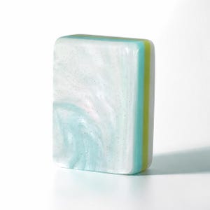 May include: A rectangular bar of soap featuring a marbled pattern of white and light blue with subtle glitter particles. The side of the soap displays distinct layered stripes in shades of light blue, lime green, and white, creating a smooth finish.
