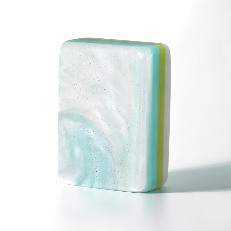 May include: A rectangular bar of soap featuring a marbled pattern of white and light blue with subtle glitter particles. The side of the soap displays distinct layered stripes in shades of light blue, lime green, and white, creating a smooth finish.