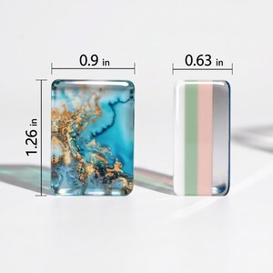 May include: Two rectangular glass cabochons. The left piece features a blue and gold marbled pattern measuring 0.9 inches wide by 1.26 inches high. The right piece has vertical stripes of white, sage green, and pale pink, measuring 0.63 inches wide.