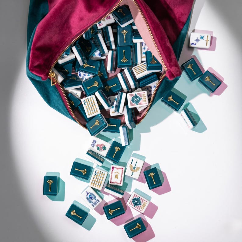 May include: A velvet pouch, in teal and magenta, spills out a collection of mahjong tiles. The tiles are teal with gold accents and white faces, some displaying symbols and numbers. The game pieces are scattered on a white surface.