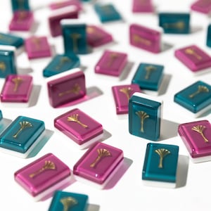 May include: A collection of colorful Mahjong tiles in shades of teal, magenta, and burgundy. Each tile features a gold-colored emblem and a white border. The tiles are scattered on a white surface, creating a visually appealing pattern.