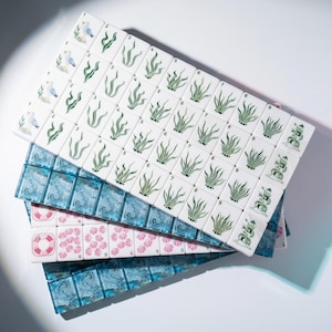 May include: A set of rectangular mahjong tiles featuring nautical designs. The white tiles display green seaweed, seagulls, and lighthouses, while others show pink seashells and life preservers. Some tiles are translucent blue with a marbled pattern.