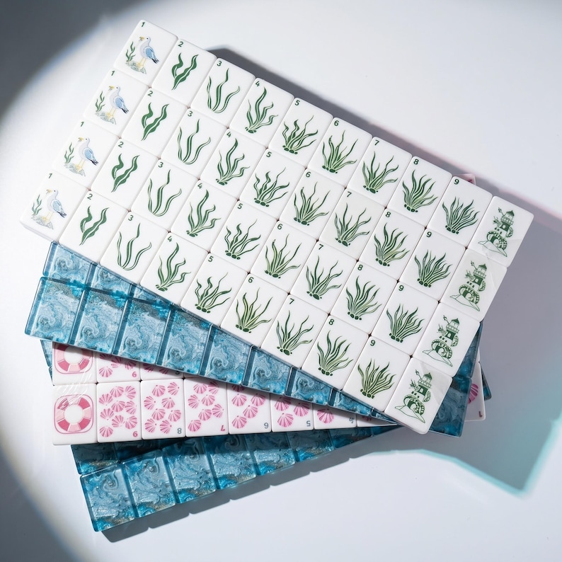 May include: A set of rectangular mahjong tiles featuring nautical designs. The white tiles display green seaweed, seagulls, and lighthouses, while others show pink seashells and life preservers. Some tiles are translucent blue with a marbled pattern.