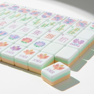 May include: A set of rectangular Mahjong tiles with a white top surface featuring colorful floral illustrations, Chinese characters, and the word JOKER. Each tile has a layered side profile with pastel green and peach bands beneath the white face.