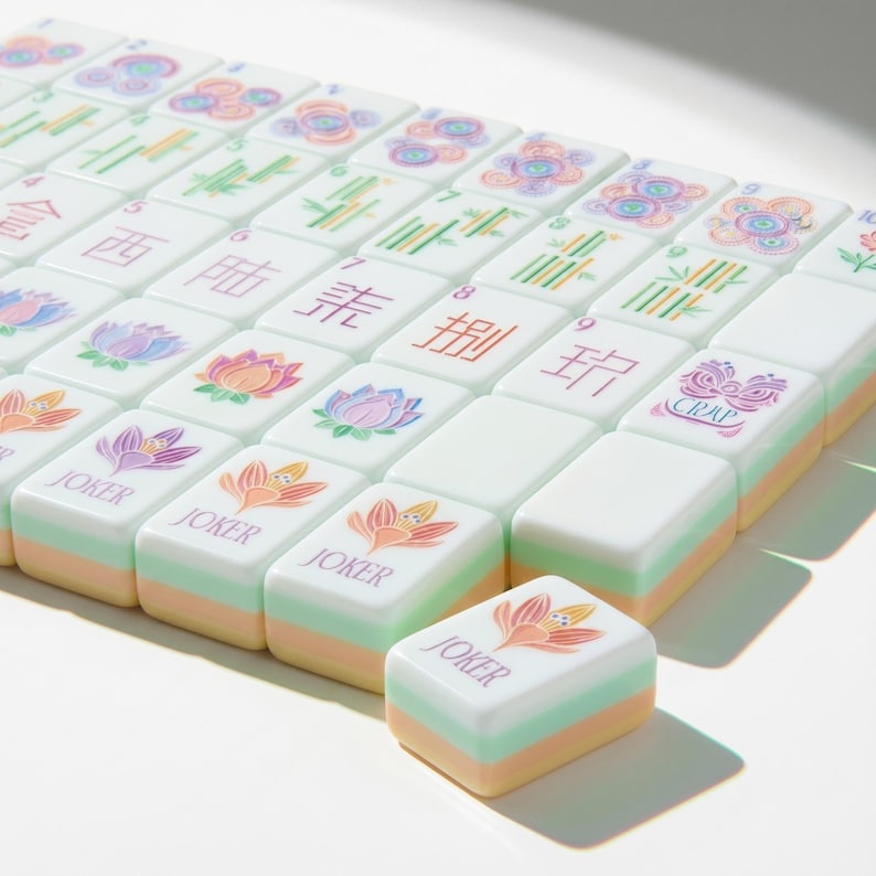 May include: A set of rectangular Mahjong tiles with a white top surface featuring colorful floral illustrations, Chinese characters, and the word JOKER. Each tile has a layered side profile with pastel green and peach bands beneath the white face.