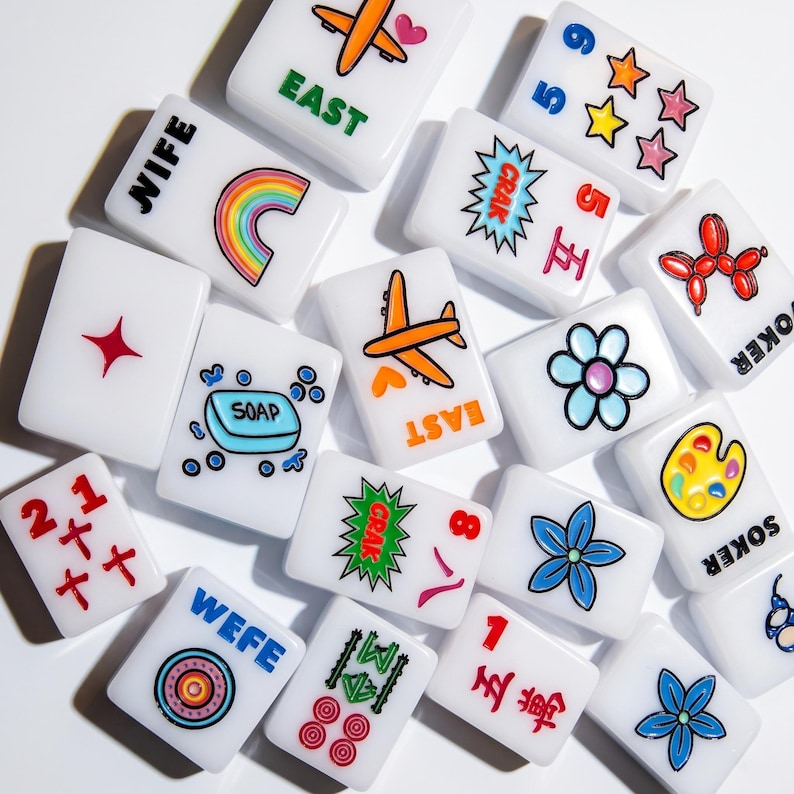 May include: A collection of white rectangular mahjong tiles featuring colorful graphic designs including a rainbow, a soap bar, airplanes, flowers, stars, a balloon dog, an artist palette, and various text labels like EAST, NIFE, and CRAK.