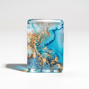 May include: A rectangular, clear resin paperweight featuring an abstract, swirling pattern of deep turquoise, light blue, and metallic gold leaf accents. The smooth, polished surface creates a glossy finish with depth and visible internal layers.