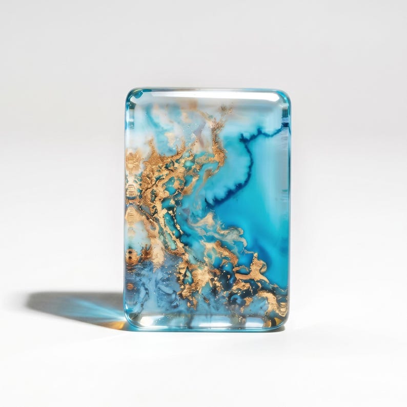 May include: A rectangular, clear resin paperweight featuring an abstract, swirling pattern of deep turquoise, light blue, and metallic gold leaf accents. The smooth, polished surface creates a glossy finish with depth and visible internal layers.