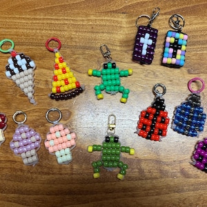 May include: A collection of twelve beaded keychains featuring various shapes including an ice cream cone, a pizza slice, two frogs, two mushrooms, two ladybugs, two cross designs, and two patterned squares, all attached to metal key rings.