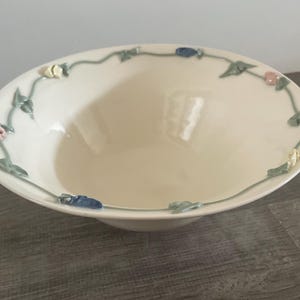 May include: A cream-colored ceramic bowl featuring a delicate, raised relief vine pattern along the inner rim. The design includes small, sculpted flowers in shades of pink, yellow, and blue, connected by thin, sage green vines and leaves.