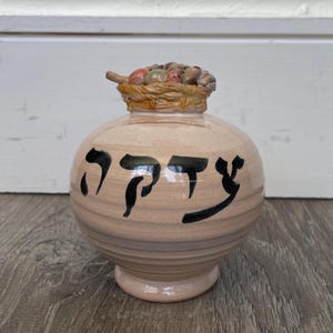 May include: A light beige ceramic jar with a rounded body and a pedestal base. The top features a sculpted basket filled with small, multi-colored fruit. Black Hebrew text is painted across the front of the jar, which has a smooth, glossy finish.