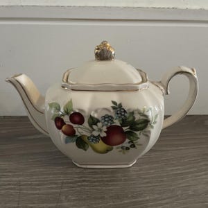 May include: A cream-colored ceramic teapot with a scalloped body and a gold-toned finial on the lid. The teapot features a colorful botanical illustration of red cherries, a yellow pear, a red apple, and blue berries with green leaves.