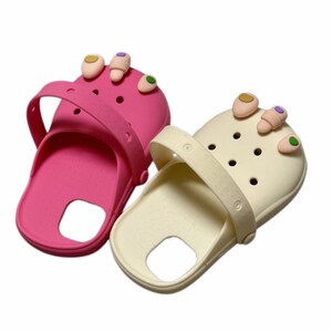 May include: A pair of slip-on clogs, one in bright pink and one in cream, each featuring a movable heel strap and a series of ventilation holes. Attached to the front of each clog are three-dimensional charms shaped like toes with painted nails.