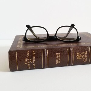 May include: A brown leather-bound book titled "The Confessions of Nat Turner" by William Styron. The book is part of The Franklin Library. A pair of black eyeglasses are resting on top of the book.