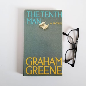 May include: A book titled "THE TENTH MAN A NOVEL" by Graham Greene. The book has a teal cover with teal and yellow lettering. A small paper origami boat is attached to the cover. Black eyeglasses rest beside the book.