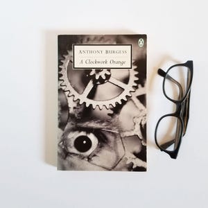 May include: A copy of Anthony Burgess's "A Clockwork Orange" with a black and white cover featuring an eye and gears. Black-framed spectacles are positioned next to the book on a white surface.