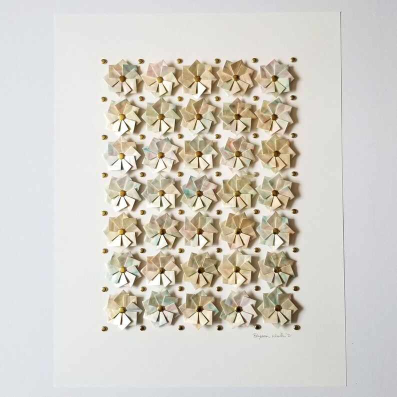 an artwork with a white background and a grid of 35 small origami flowers in shades of beige, pink, and blue. The flowers are arranged in a grid pattern and are accented with gold tone metal pins. The artwork is signed in the bottom right corner.