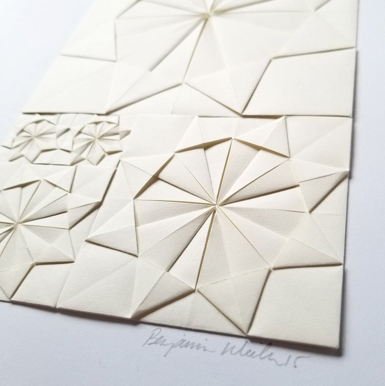 an origami artwork of ivory coloured paper on a white background. The artwork is a geometric design made of folded paper squares. The design is made up of several different shapes, including circles, squares, and triangles. The design is symmetrical and has a radial pattern. The artwork is signed in the bottom right corner.