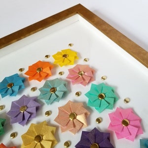 a corner detail of a gold framed artwork featuring a rainbow of origami paper flowers with gold centers. The flowers are arranged in a grid pattern on a white background.