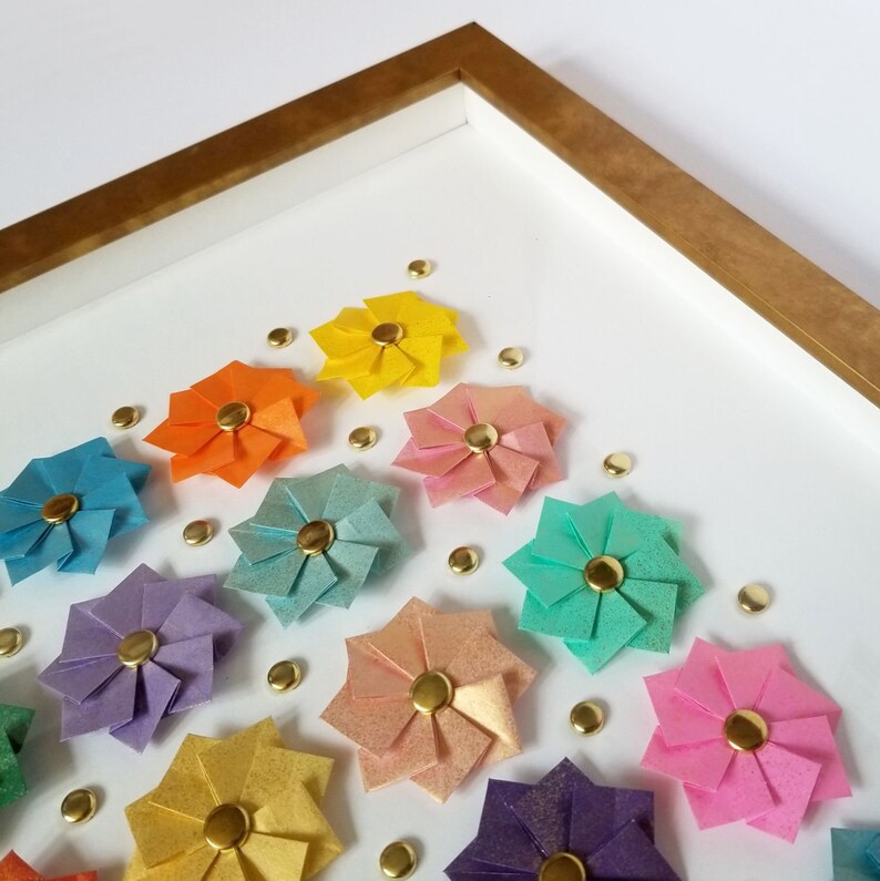 a corner detail of a gold framed artwork featuring a rainbow of origami paper flowers with gold centers. The flowers are arranged in a grid pattern on a white background.