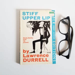 May include: A vintage paperback book titled "Stiff Upper Lip" by Lawrence Durrell. The cover features a black and white illustration of a man in a tuxedo sitting on a stool with a palm tree in the background. The book is part of the Coles Paper Covered Editions series.