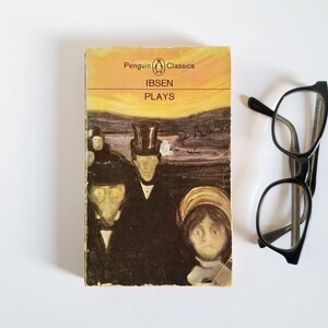 May include: A Penguin Classics paperback book with a yellow, orange, and purple cover featuring a painting of three people. The title of the book is "IBSEN PLAYS".