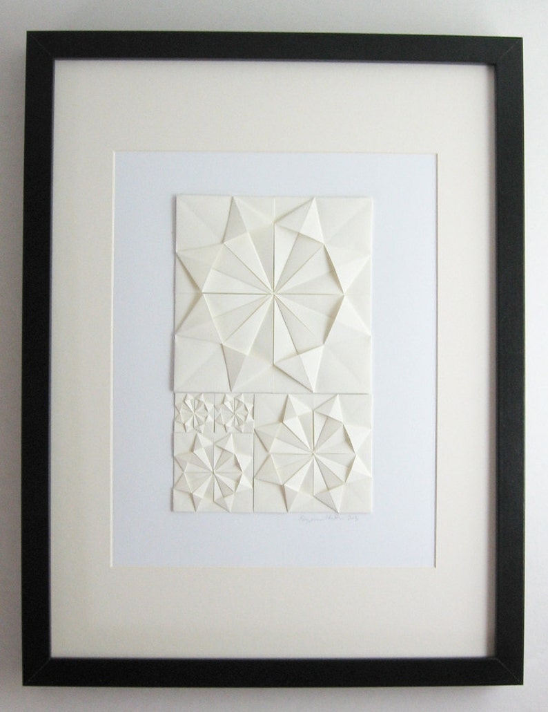 an origami artwork of ivory coloured paper on a white background. The artwork is a geometric design made of folded paper squares. The design is made up of several different shapes, including circles, squares, and triangles. The design is symmetrical and has a radial pattern. The artwork is signed in the bottom right corner. The artwork is shown in a black frame with mat, not included.
