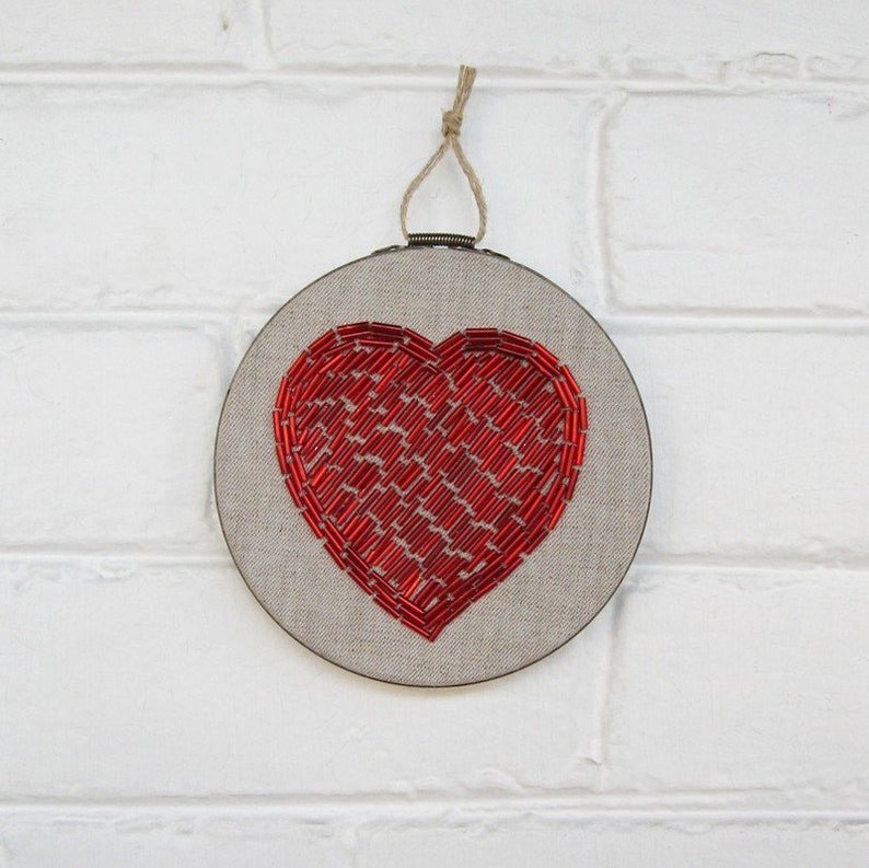 May include: A red beaded heart embroidered on a round piece of beige linen fabric, framed in a wooden hoop.