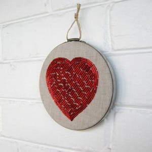 May include: A red beaded heart embroidered on a beige linen fabric stretched over a round wooden embroidery hoop.