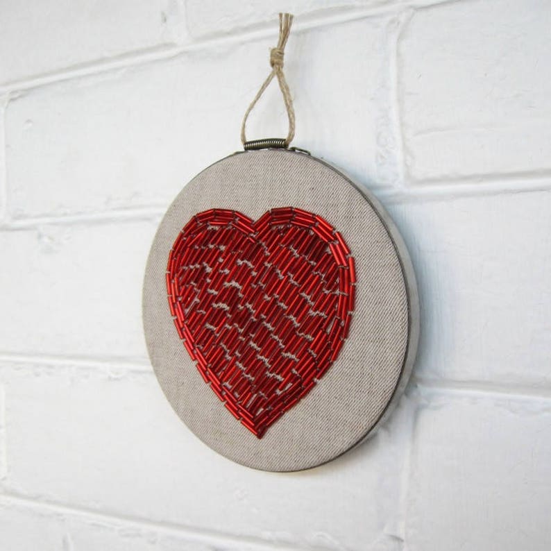 May include: A red beaded heart embroidered on a beige linen fabric stretched over a round wooden embroidery hoop.